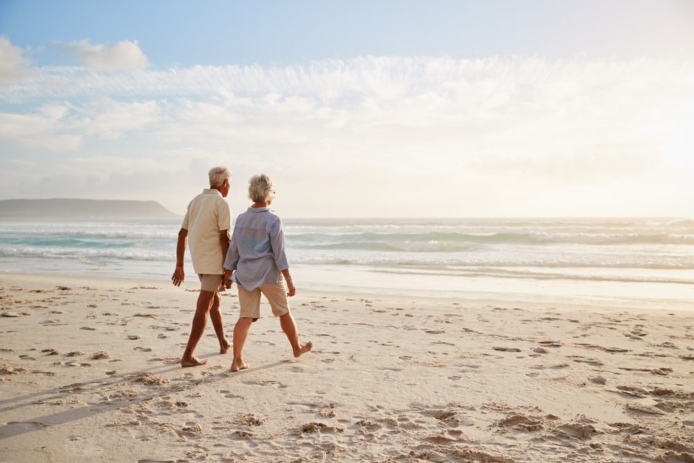 Rear,View,Of,Senior,Couple,Walking,Along,Beach,Hand,In